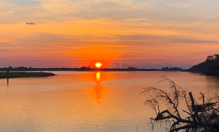 A sunrise casts an orange glow over a cloudy sky and a tranquil lake, with trees lining the shore.