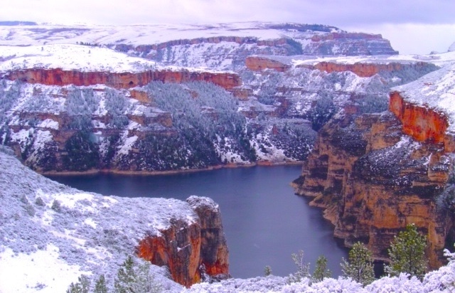 Snow-covered cliffs of a deep canyon with a lake on the canyon floor, under a cloudy sky.