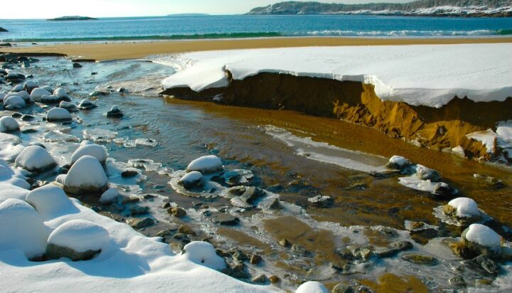 A river mouth at the beach of a larger body of water, with broken snowbanks along the river's edge and ice attached to rocks in the river.